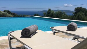 Une vue sur la piscine, avec deux chaises longues et des serviettes, donnant sur une mer d’un bleu limpide et sur des collines lointaines.