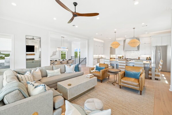 A spacious living room with a sofa, armchairs, and a coffee table. There is a kitchen area with modern appliances and pendant lighting.