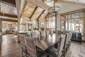 Un vaste espace dédié à la salle à manger, avec une longue table en bois et des chaises à motifs, un plafond haut avec des poutres apparentes et de grandes fenêtres offrant une vue sur l’extérieur.