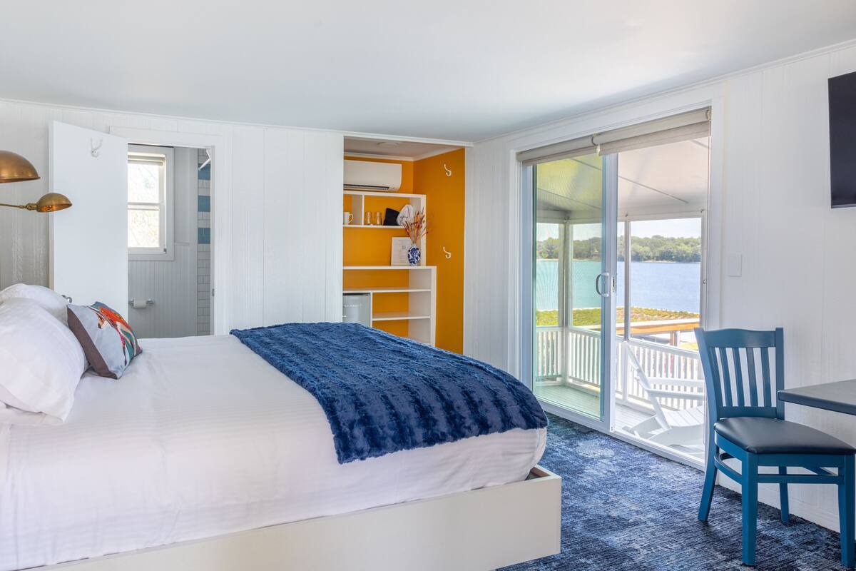 Ein Schlafzimmer mit Bett, Schreibtisch, Stuhl und einem Balkon mit Blick auf das Wasser. Ein offenes Kleiderschrank mit Regalen.