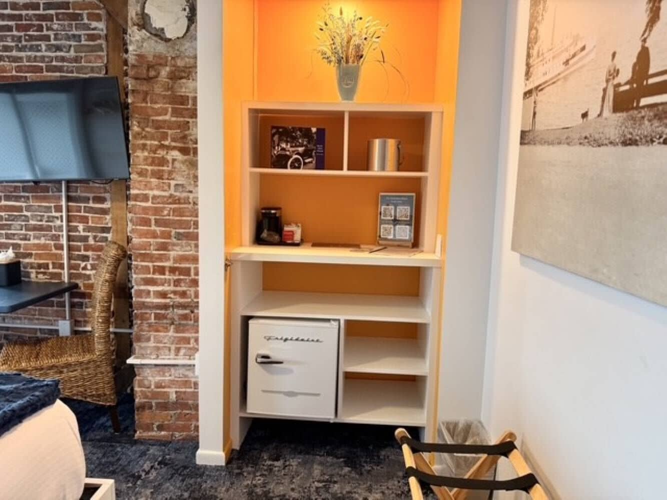 Une pièce avec un mur en briques, une étagère blanche avec un petit réfrigérateur, une chaise en osier et un tableau encadré accroché au mur.