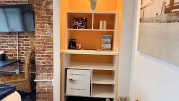 Une pièce avec un mur en briques, une étagère blanche avec un petit réfrigérateur, une chaise en osier et un tableau encadré accroché au mur.