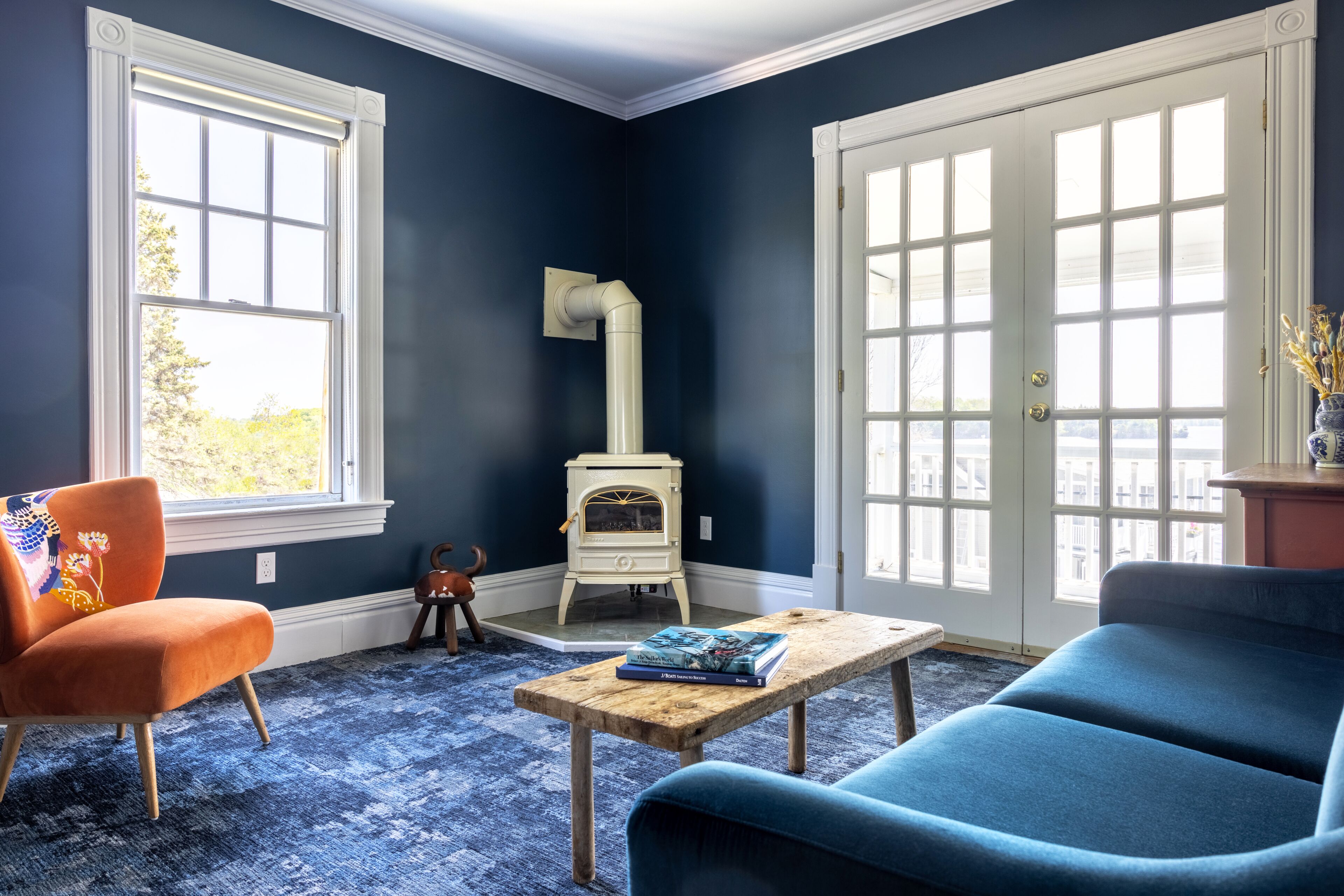 A room with a blue wall, a white door, a blue sofa, an orange chair, a wooden table, and a white stove.
