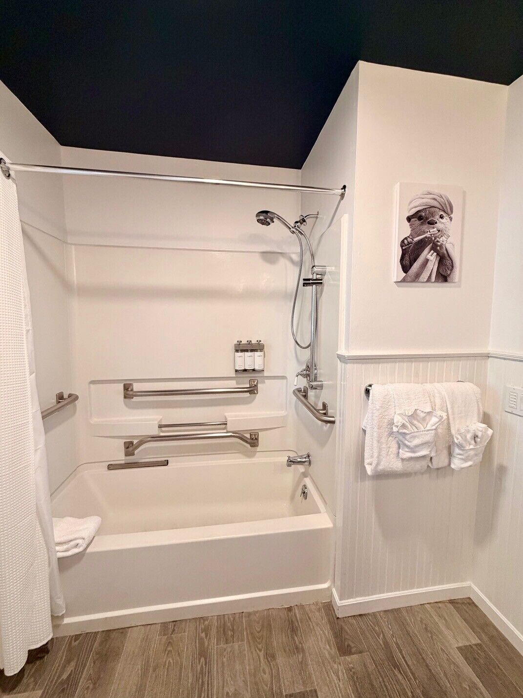A modern bathroom with a bathtub, shower, and handrails. There is a towel rack with towels and a framed picture on the wall.