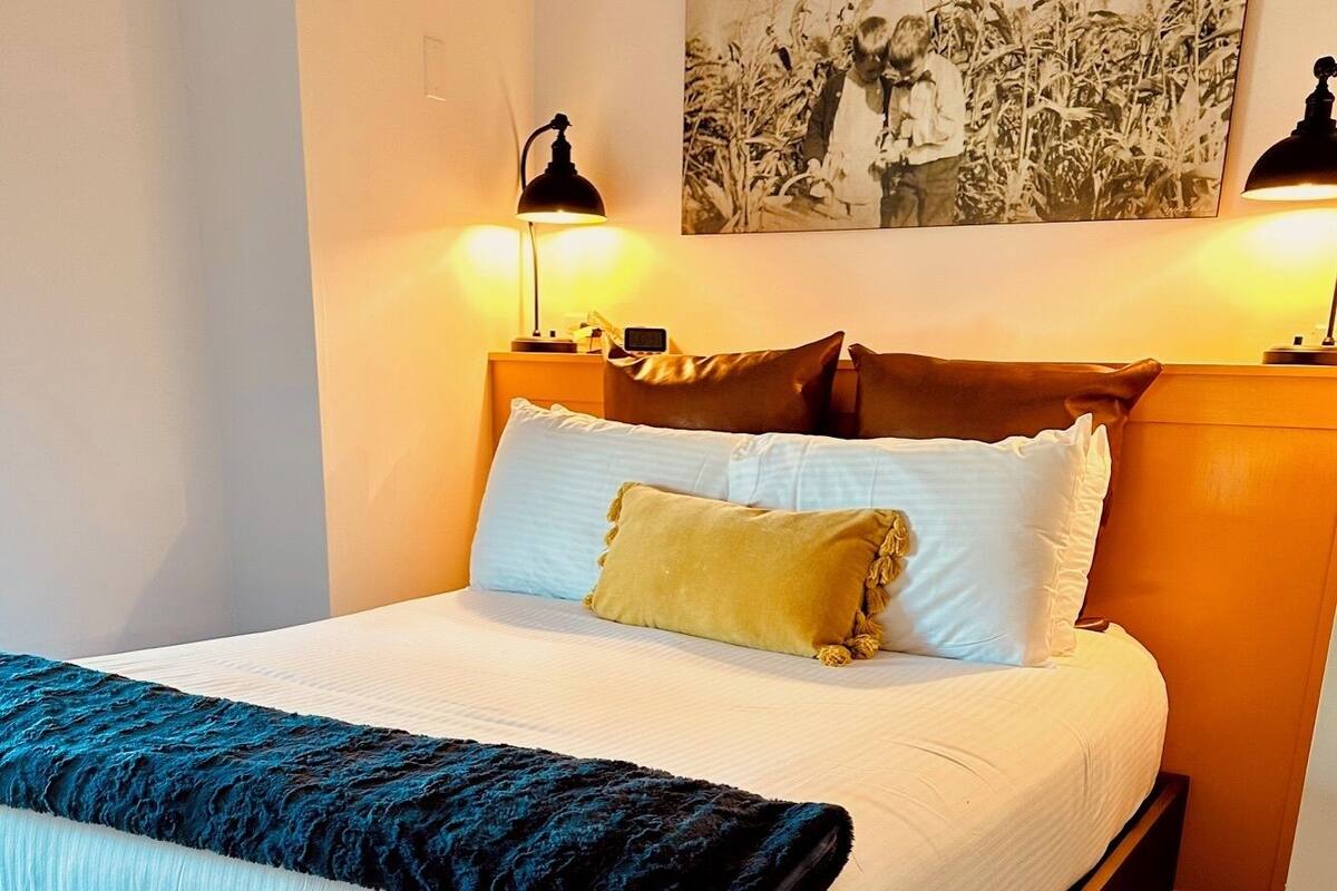 Ein Bett mit weißer Bettwäsche, einer blauen und schwarzen Decke und einem braunen Kopfteil. Zwei Nachttischlampen und ein gerahmtes Bild sind an der Wand zu sehen.