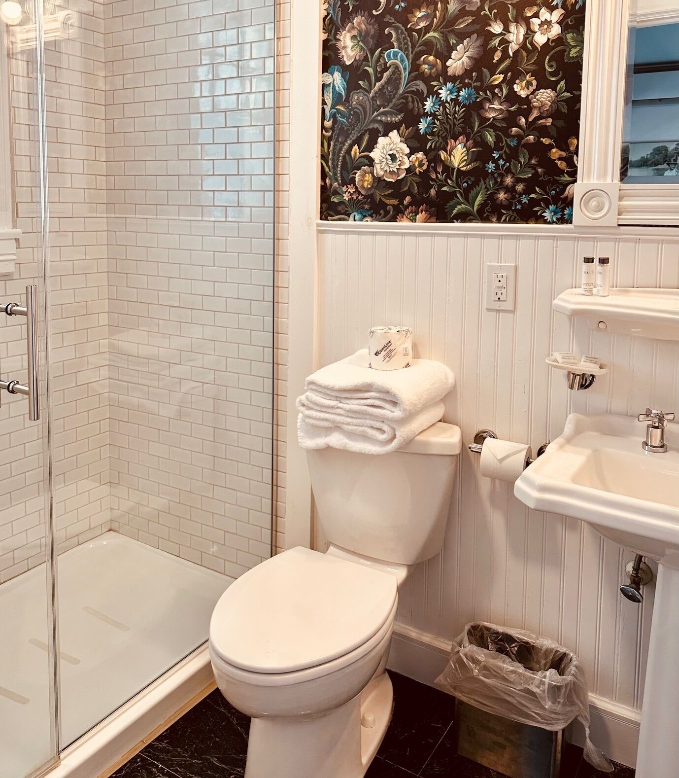 A bathroom with a white toilet, a shower enclosure, a sink, and a decorative floral wallpaper.