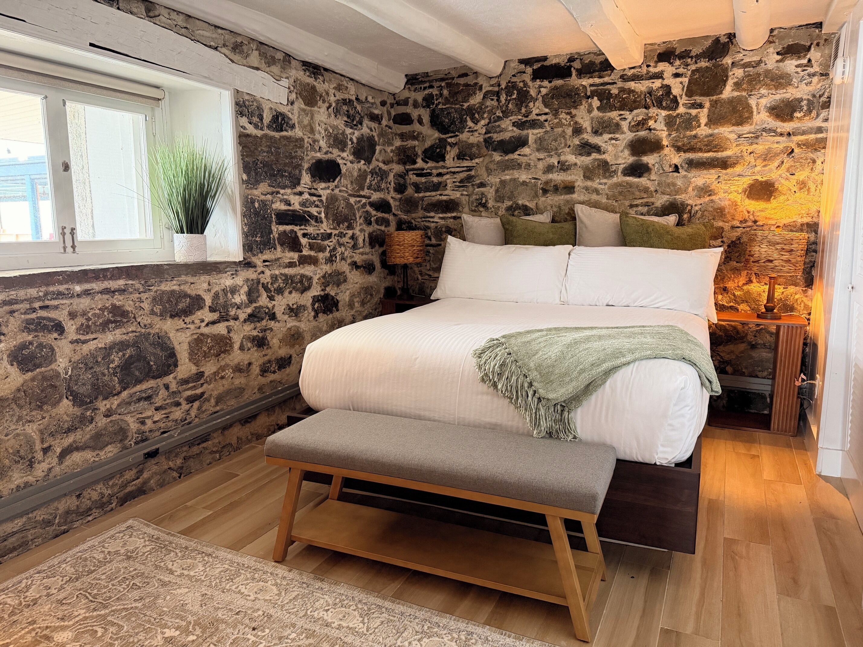 Une chambre avec un mur en pierre, un lit avec une literie blanche, un banc, une table de chevet avec une lampe et une fenêtre agrémentée d’une plante.