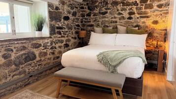 Une chambre avec un mur en pierre, un lit avec une literie blanche, un banc, une table de chevet avec une lampe et une fenêtre agrémentée d’une plante.
