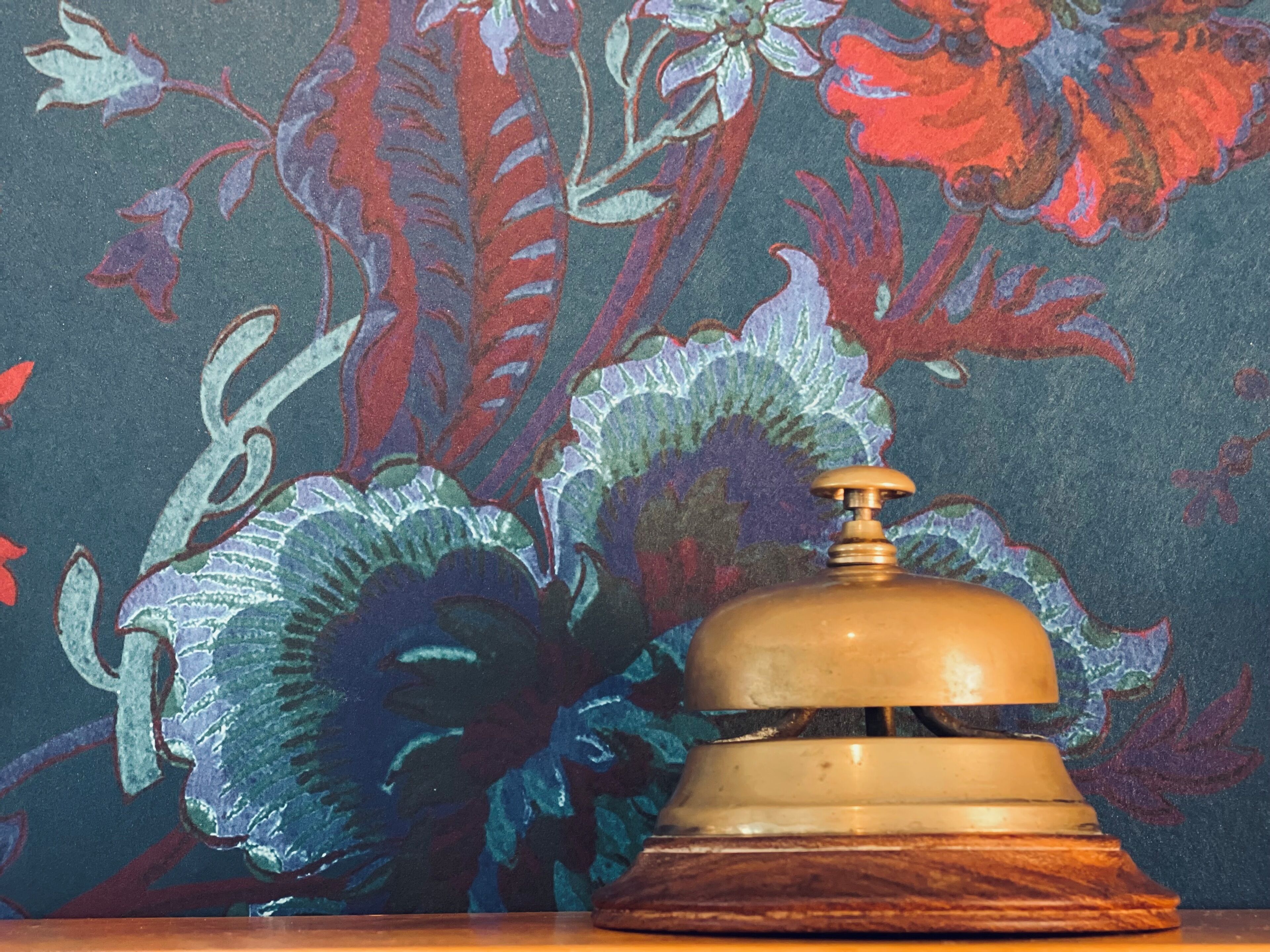 Une cloche en laiton, posée sur un socle en bois, devant un mur orné de motifs floraux.