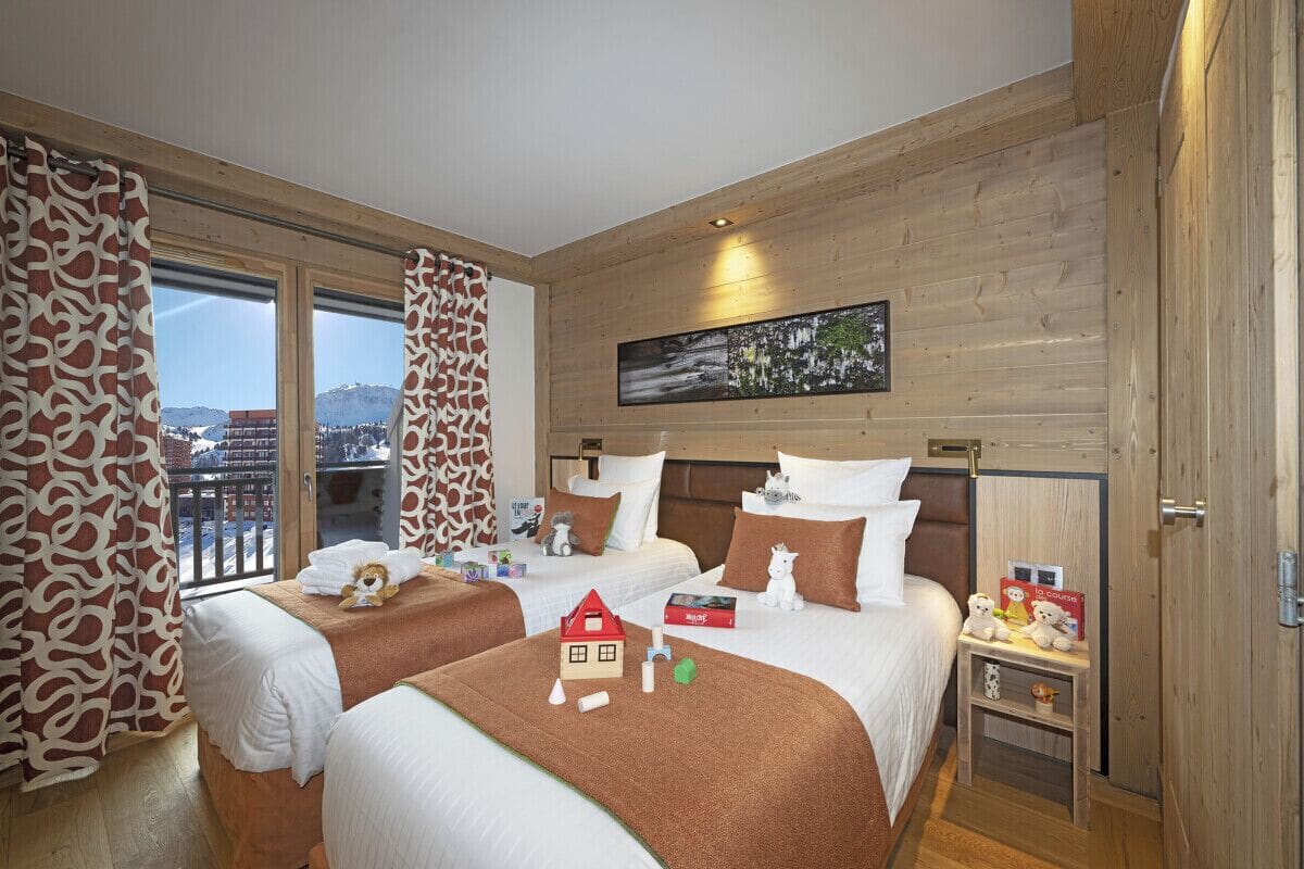 A hotel room with two beds, wooden walls, a large window with curtains, a painting above the beds, and a small table with items.