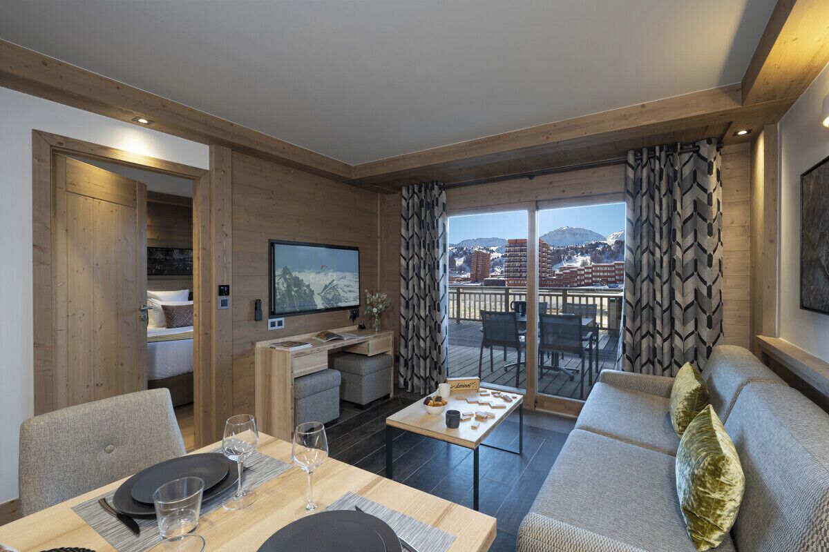 A modern living room with a dining area, a sofa, and a television. There is a balcony with a view of buildings and mountains.