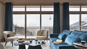 A modern living room with a sofa, armchairs, a coffee table, and a view of snow-covered mountains through large windows. - MGM Hôtels & Résidences – Résidence Manaka (La Plagne-Tarentaise)