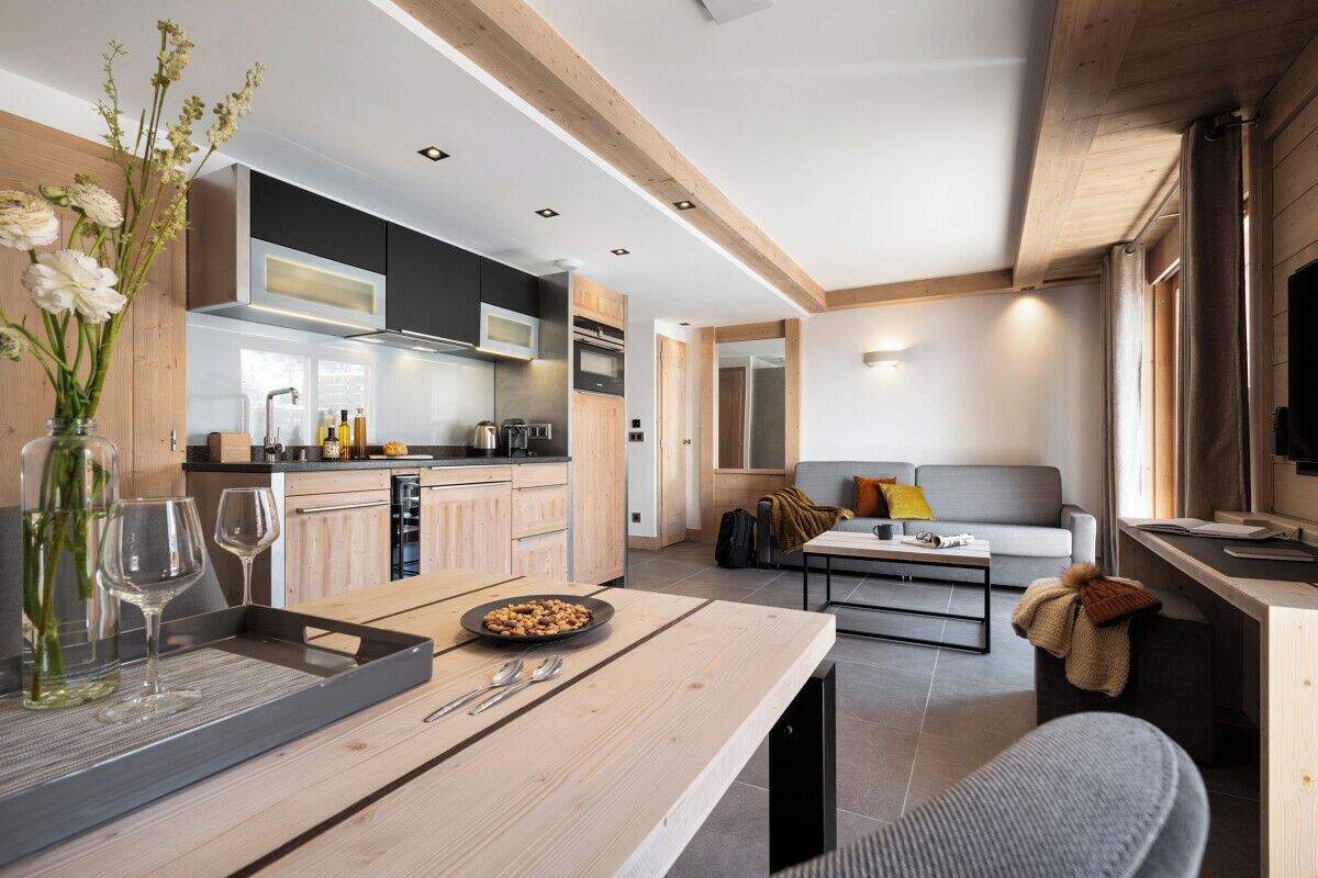 A modern kitchen with wooden cabinets, a dining area with a wooden table, and a living room with a sofa and a television.