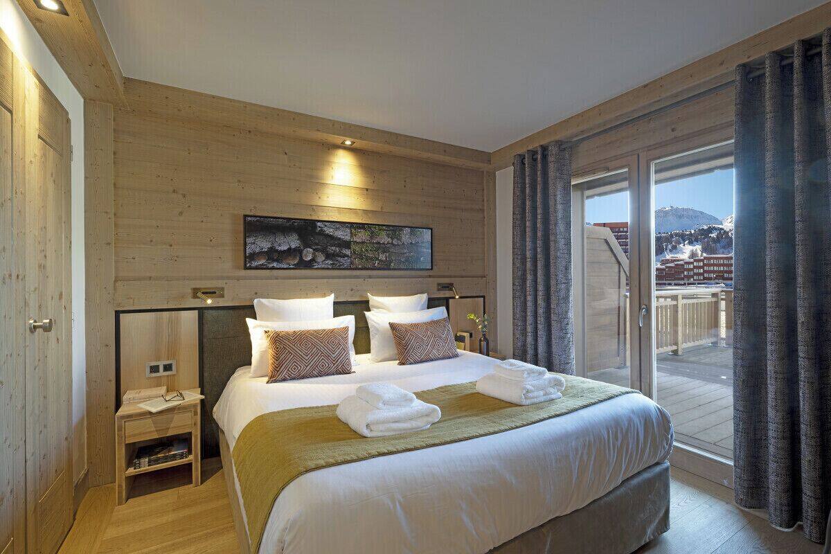 A hotel room with a large bed, wooden panel walls, a painting above the bed, and a view of a snowy landscape through the window.