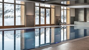 A large indoor swimming pool with clear glass walls, wooden beams, and a view of a snowy mountain landscape. - MGM Hôtels & Résidences – Résidence Manaka (La Plagne-Tarentaise)