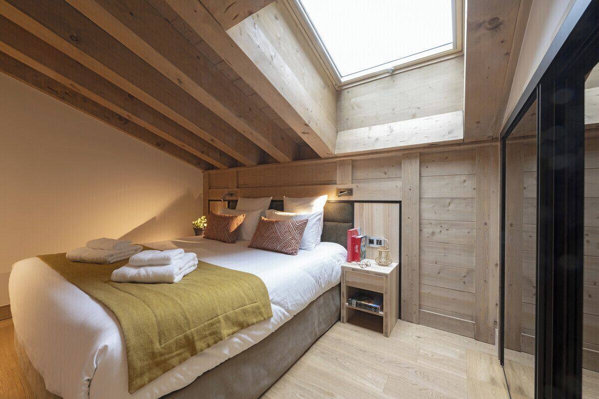 A bedroom with a skylight, a bed with pillows and a blanket, a bedside table with books and a lamp, and wooden walls.