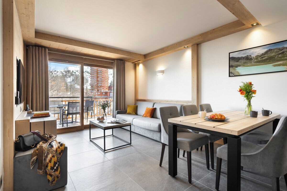 A modern living room with a sofa, dining table, and chairs. There is a balcony with a view of buildings and a lake.
