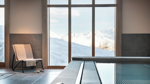 A spa area with a chair, a massage table, and a view of snow-covered mountains through large windows. - MGM Hôtels & Résidences – Résidence Manaka (La Plagne-Tarentaise)