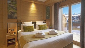 A wooden-paneled bedroom with a large bed, two pillows, a nightstand, and a view of mountains through a sliding glass door. - MGM Hôtels & Résidences – Résidence Manaka (La Plagne-Tarentaise)
