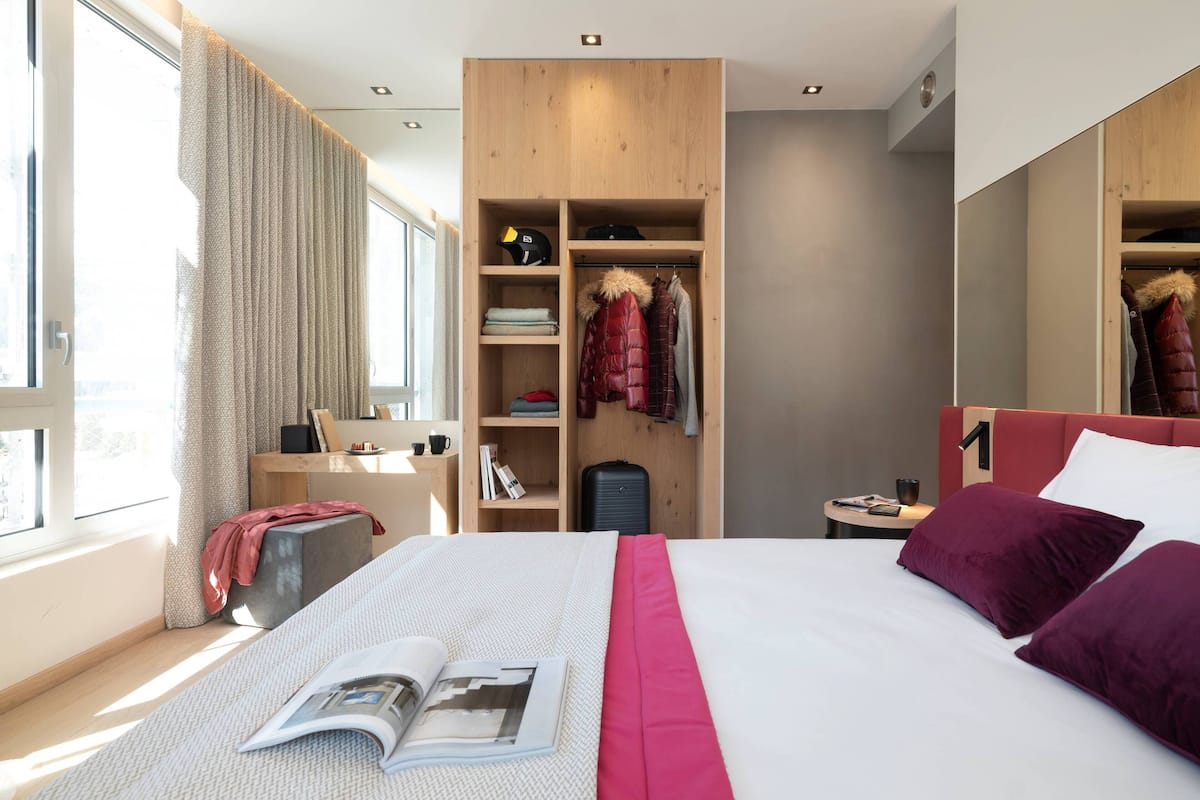 Moderni hotellihuone, jossa on suuri sänky, puinen vaatekaappi, työpöytä ja ikkuna, jossa on verhot.