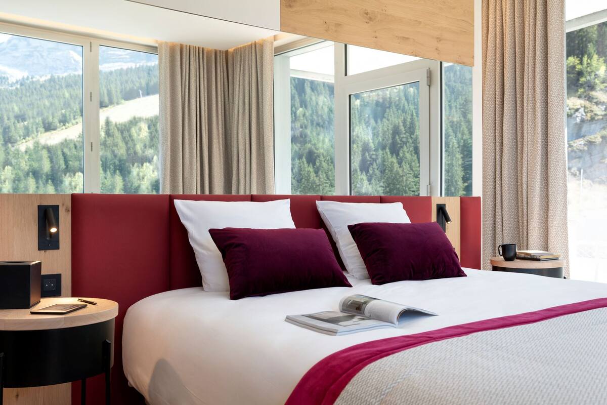 Hotellihuone, jossa on suuri sänky, punainen pääty ja näkymä metsäiselle vuoristolle.