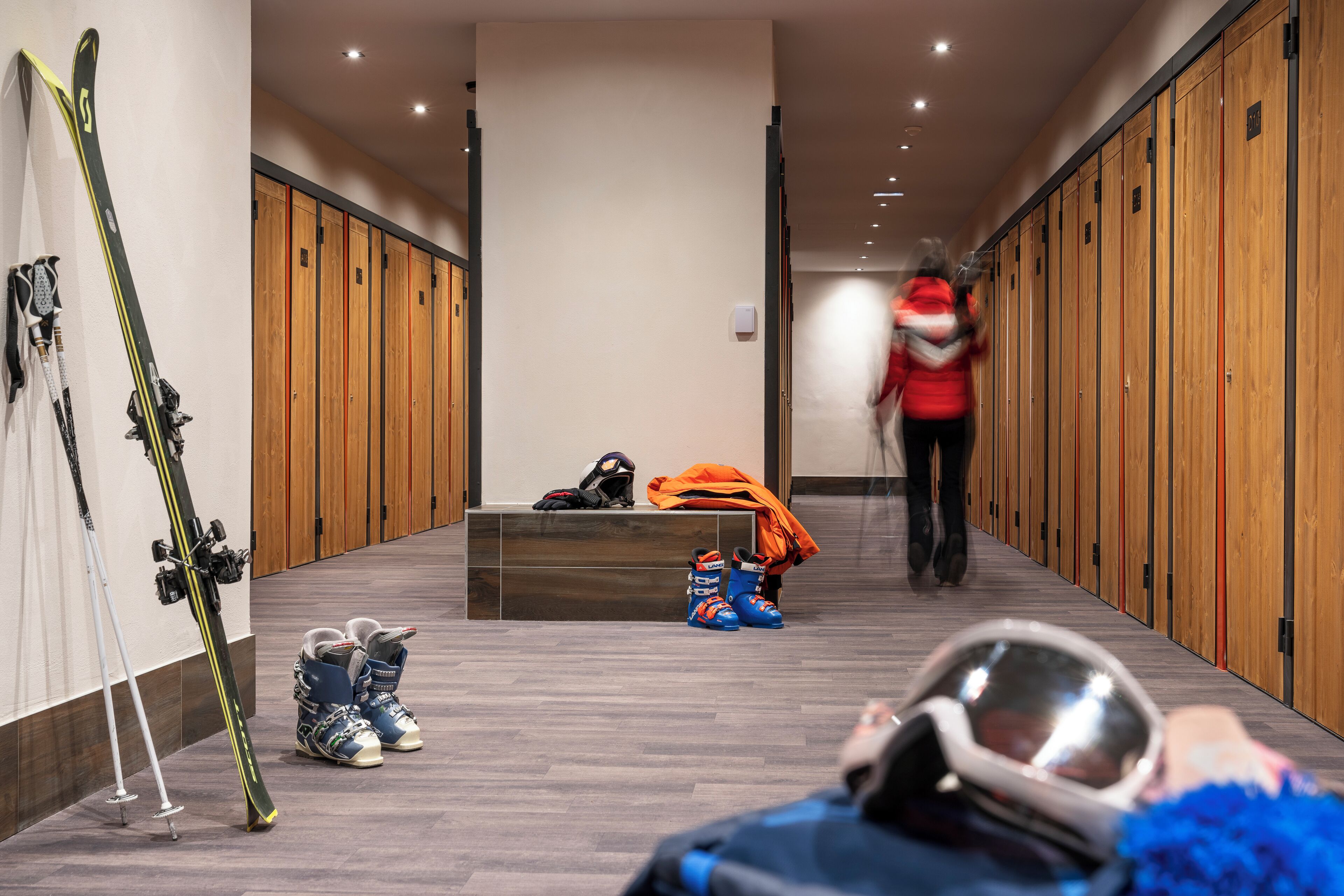 A hallway with lockers, skis leaning against the wall, and a person walking.