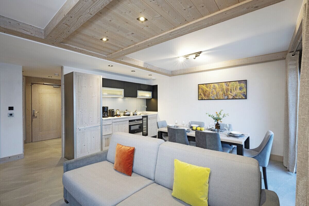 Una sala de estar moderna con un sofá gris, un comedor con mesa y sillas, una cocina con armarios blancos y un techo de madera.