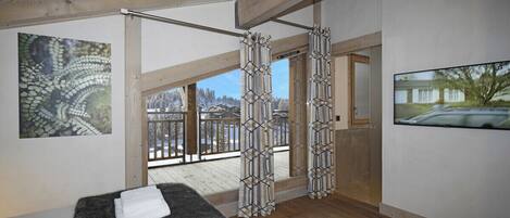 Ein Zimmer mit Holzboden, einem großen Fenster mit Blick auf eine verschneite Landschaft, einem schwarzen Teppich mit Handtüchern und einem Baumgemälde.