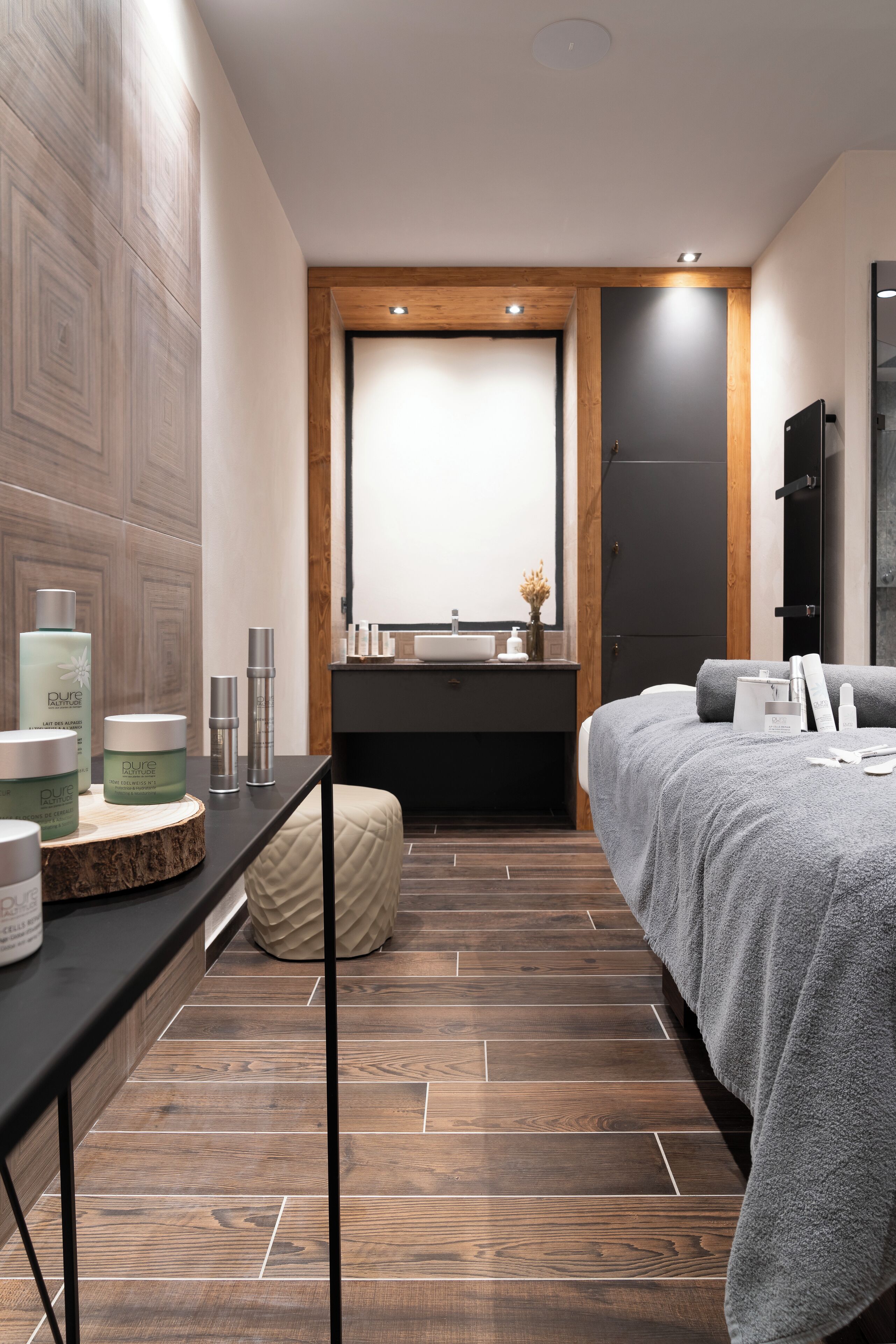 A modern bedroom with a wooden floor, a bed with grey bedding, a bedside table with toiletries, and a large mirror above the dresser.