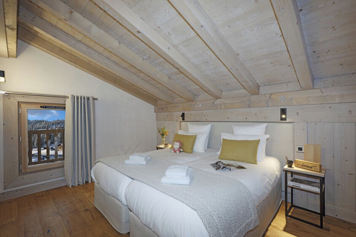 A bedroom with a wooden ceiling, a bed with white bedding, a nightstand with a lamp, and a window with curtains.