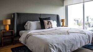 Ein modernes Schlafzimmer mit einem großen Bett, zwei Nachttischlampen, einem grauen Kopfteil und Blick auf die Stadt durch große Fenster.