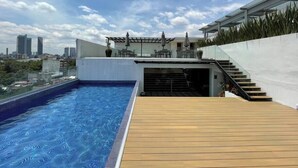 A rooftop swimming pool with a wooden deck, a covered seating area, and a cityscape view in the background. - Great location, pool, rooftop, gym, 24/7 security (Ciudad de México)