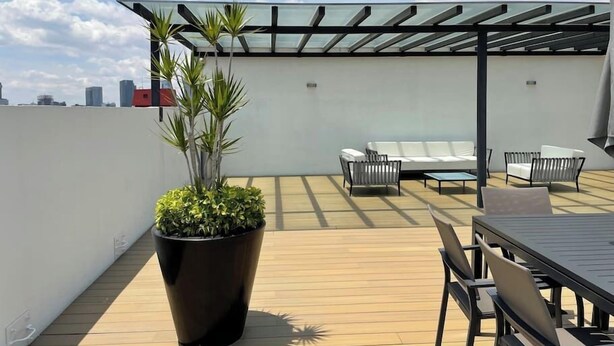 Een terras op het dak met een glazen pergola, een witte bank en een eettafel met stoelen.
