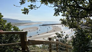 Une vue sur la côte, prise depuis un escalier en bois muni d’une barrière en corde, donnant sur une plage où se trouvent des bateaux et, au loin, une ligne de rivage.