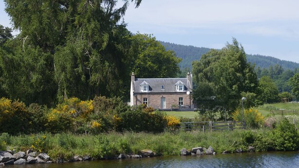 A house by a lake with a stone facade, surrounded by trees and a grassy area.