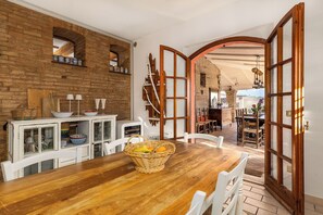 A dining area with a wooden table, white chairs, a brick wall, and an open door leading to another room.