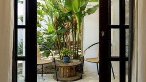 Een geopende deur die uitkomt op een klein terras met een ronde houten tafel, twee rieten stoelen en een bloempot.