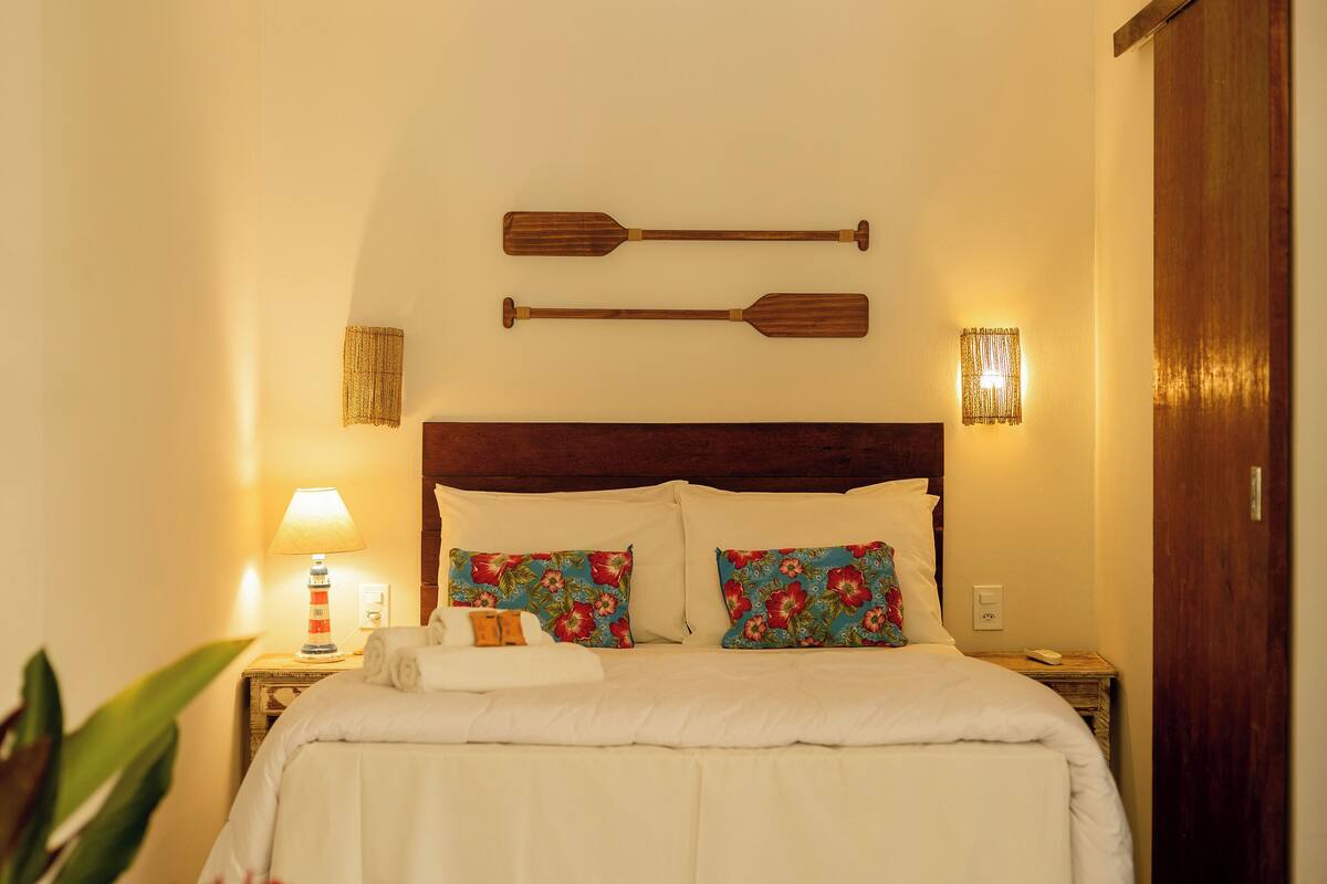 A neatly made bed with white bedding and colorful floral pillows, a wooden headboard, and two wooden paddles mounted on the wall above.