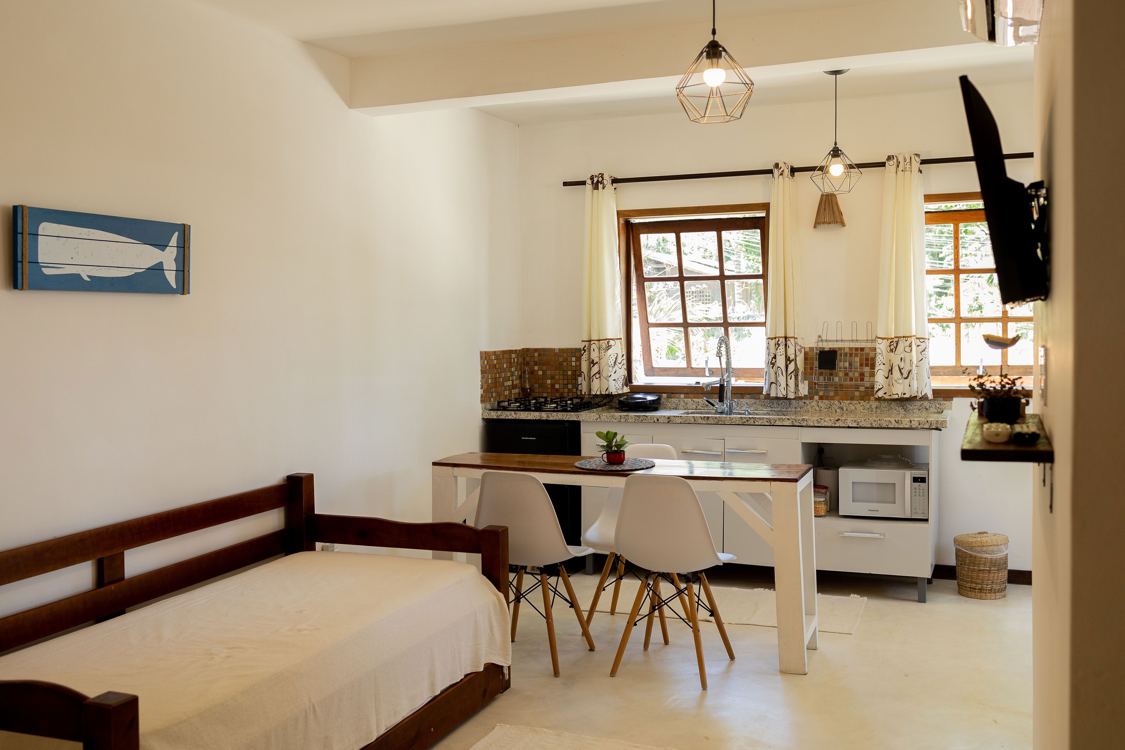 A bedroom with a wooden bed, a dining area with chairs and a table, a kitchen with a stove and microwave, and a window with curtains.