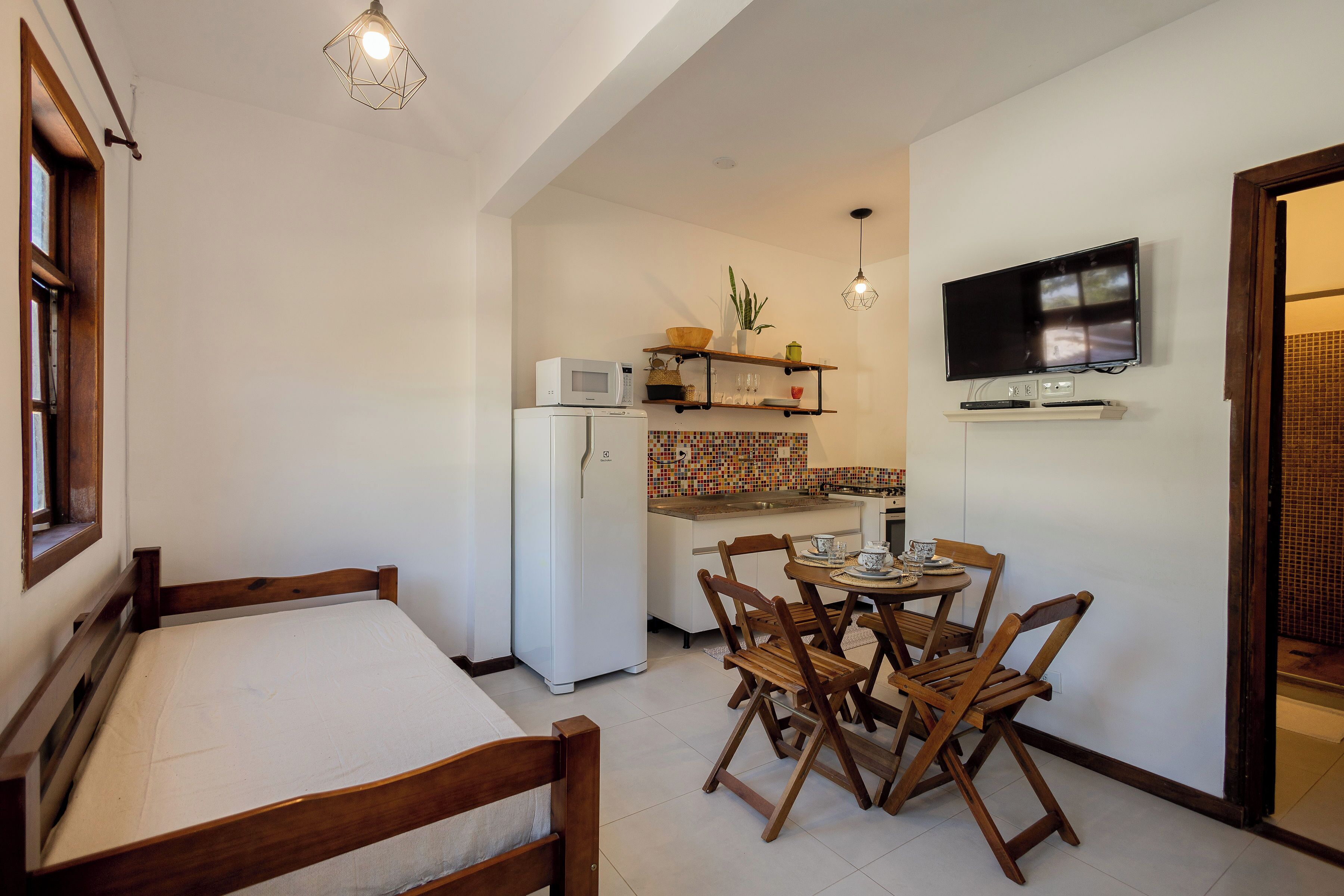 A compact kitchen with a microwave, refrigerator, and a TV mounted on the wall. There is a dining area with wooden chairs and a table.