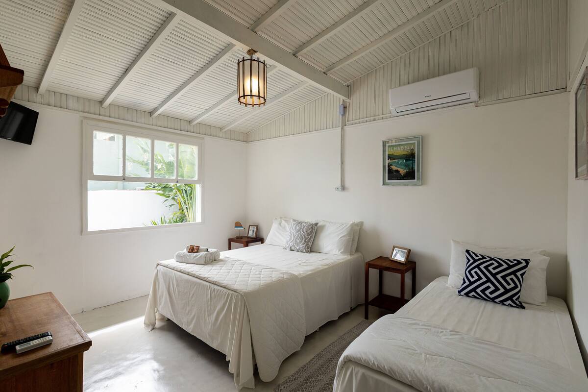 A bedroom with two beds, a ceiling fan, an air conditioning unit, a window with a view of greenery, and a mounted television.