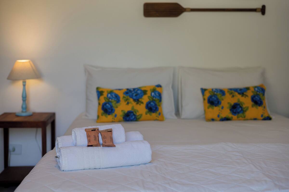 A neatly made bed with white linens and a folded towel, two pillows with a blue floral pattern, a wooden paddle wall hanging, and a bedside lamp on a wooden nightstand.