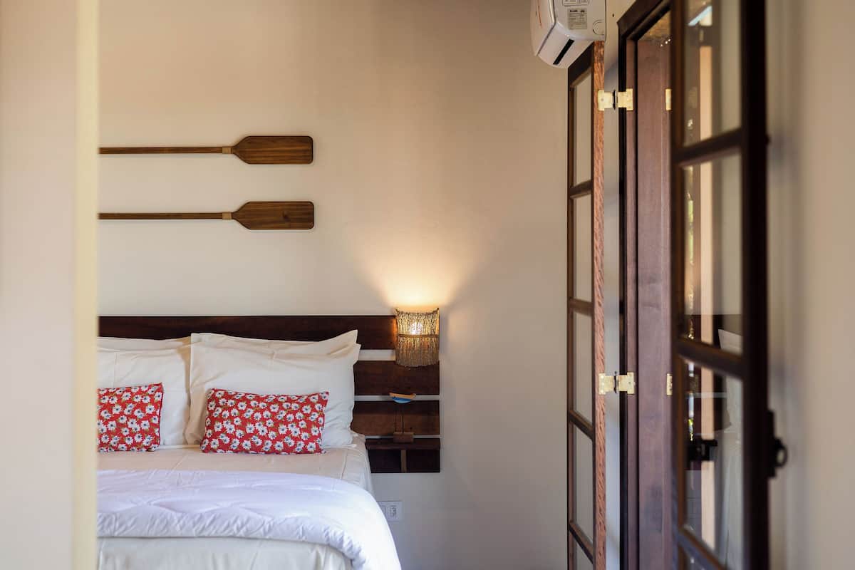 A neatly made bed with white linens and red floral pillows, a wooden headboard, and a wall-mounted light fixture.