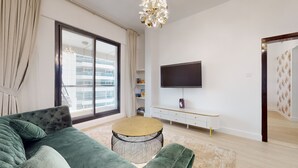Uma sala de estar moderna com um sofá verde, uma mesa de centro redonda, uma TV de tela plana na parede e uma janela grande com vista para um edifício.