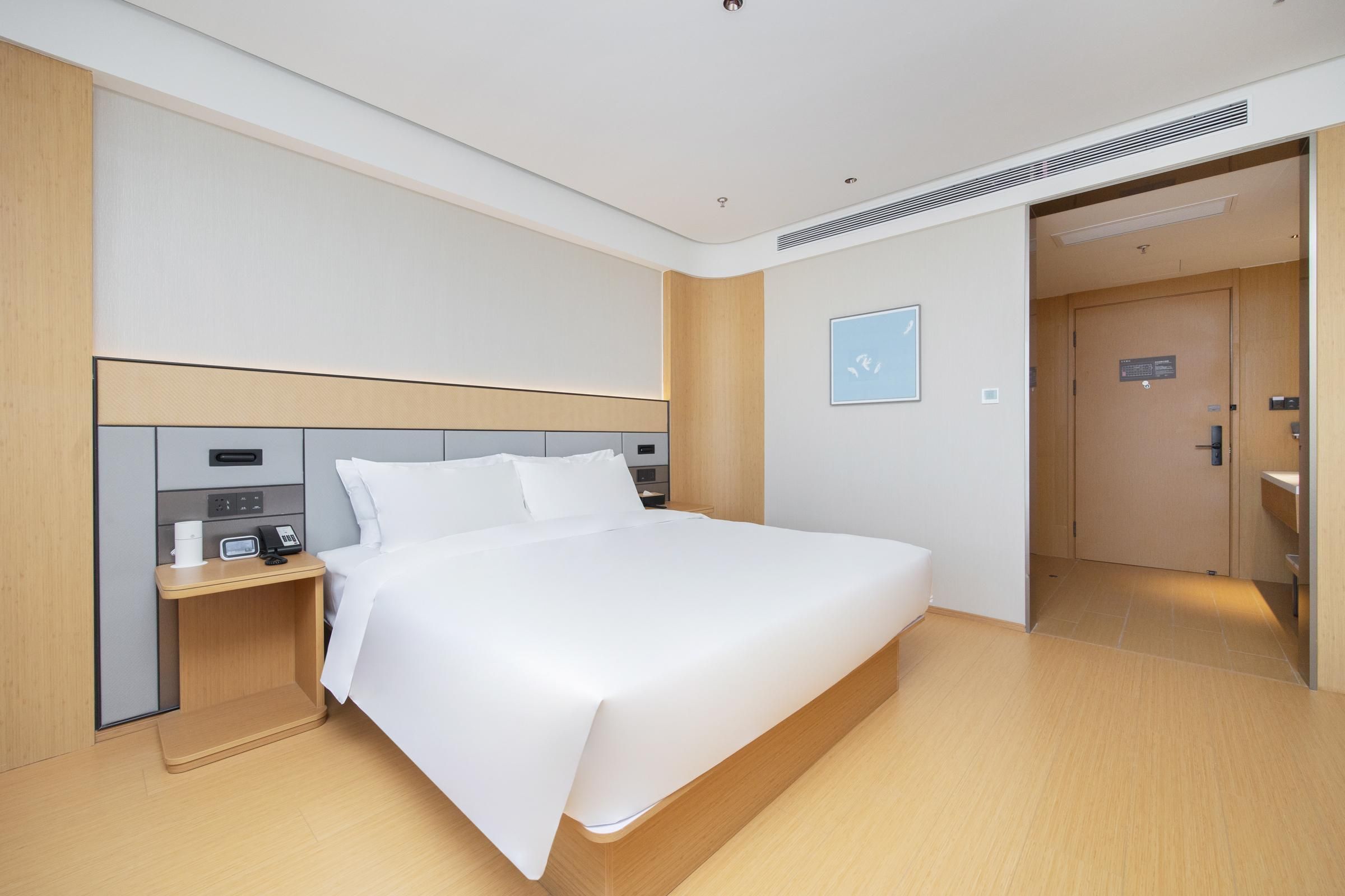 Una habitación de hotel moderna con una cama grande, una mesita de noche con teléfono y cafetera, un cabecero de madera, y una puerta que da a un baño.