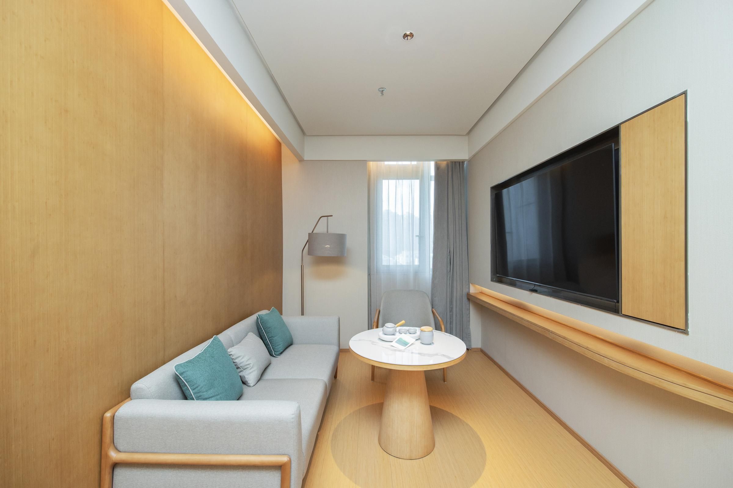 A modern hotel room with a sofa, a round table with a teapot and cups, a flat-screen TV mounted on the wall, and a window with curtains.