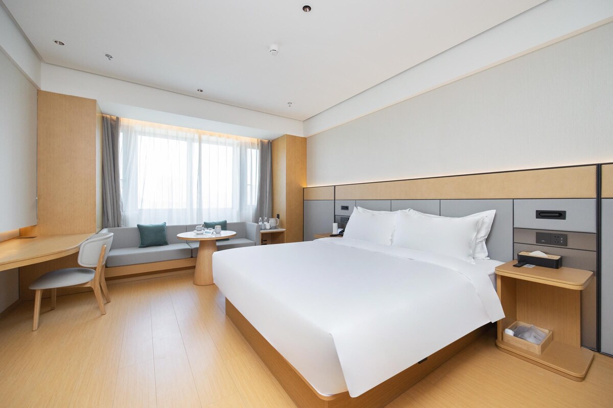 A modern hotel room with a large bed, a desk, a chair, a small table, and a window with curtains.