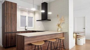 Une cuisine moderne avec un îlot central en bois, des tabourets de bar, une hotte noire et une fenêtre équipée de stores.