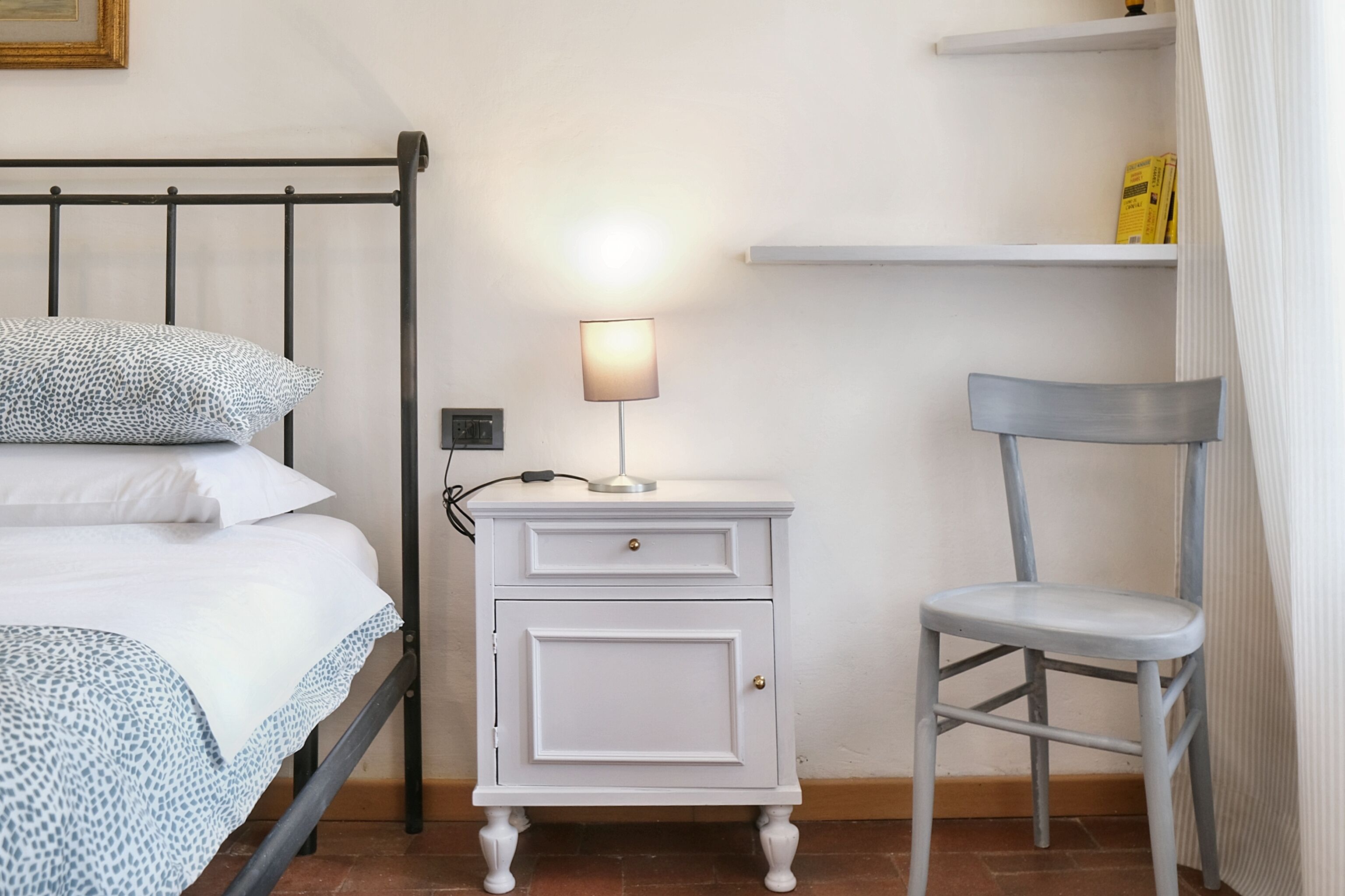 A neatly made bed with patterned bedding, a white bedside table with drawers, a lamp, and a grey chair.