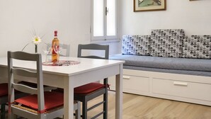 Ruokailutilassa on valkoinen pöytä, punaisilla istuintyynyillä varustetut tuolit, maljakko, jossa on yksi päivänkukka, ja pullo viiniä. Harmaa sohva, jossa on kuvioiduilla tyynyillä, on seinää vasten.